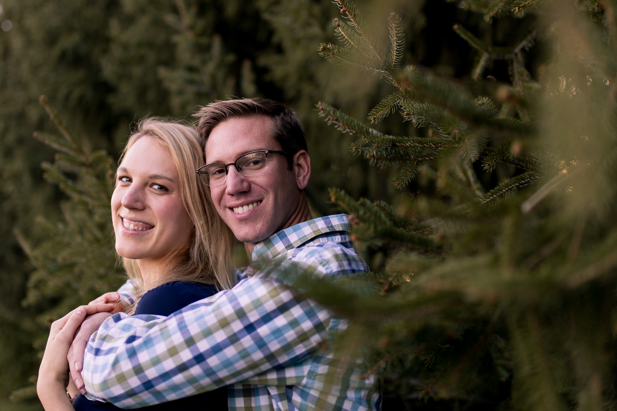 bride and groom look at camera by pine trees during winter farm engagement session in Eaton Indiana