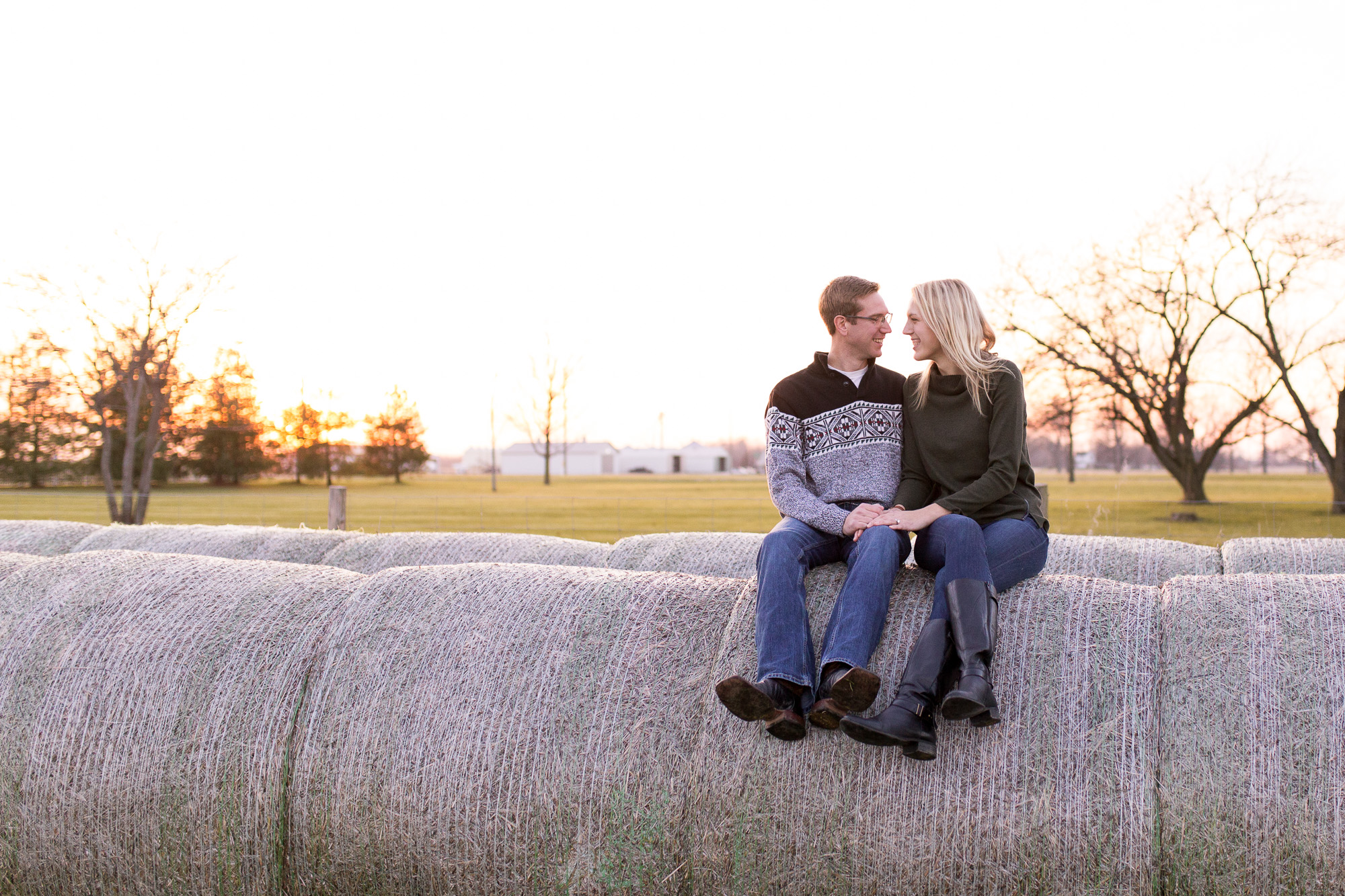 bride and groom hold hands while sitting on hay bales during Eaton Indiana winter farm engagement