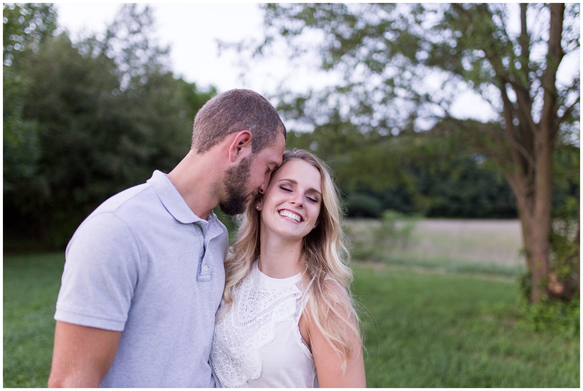 bride grins at the camera while groom nuzzles into her hair during Fort Wayne engagement session at Metea County Park